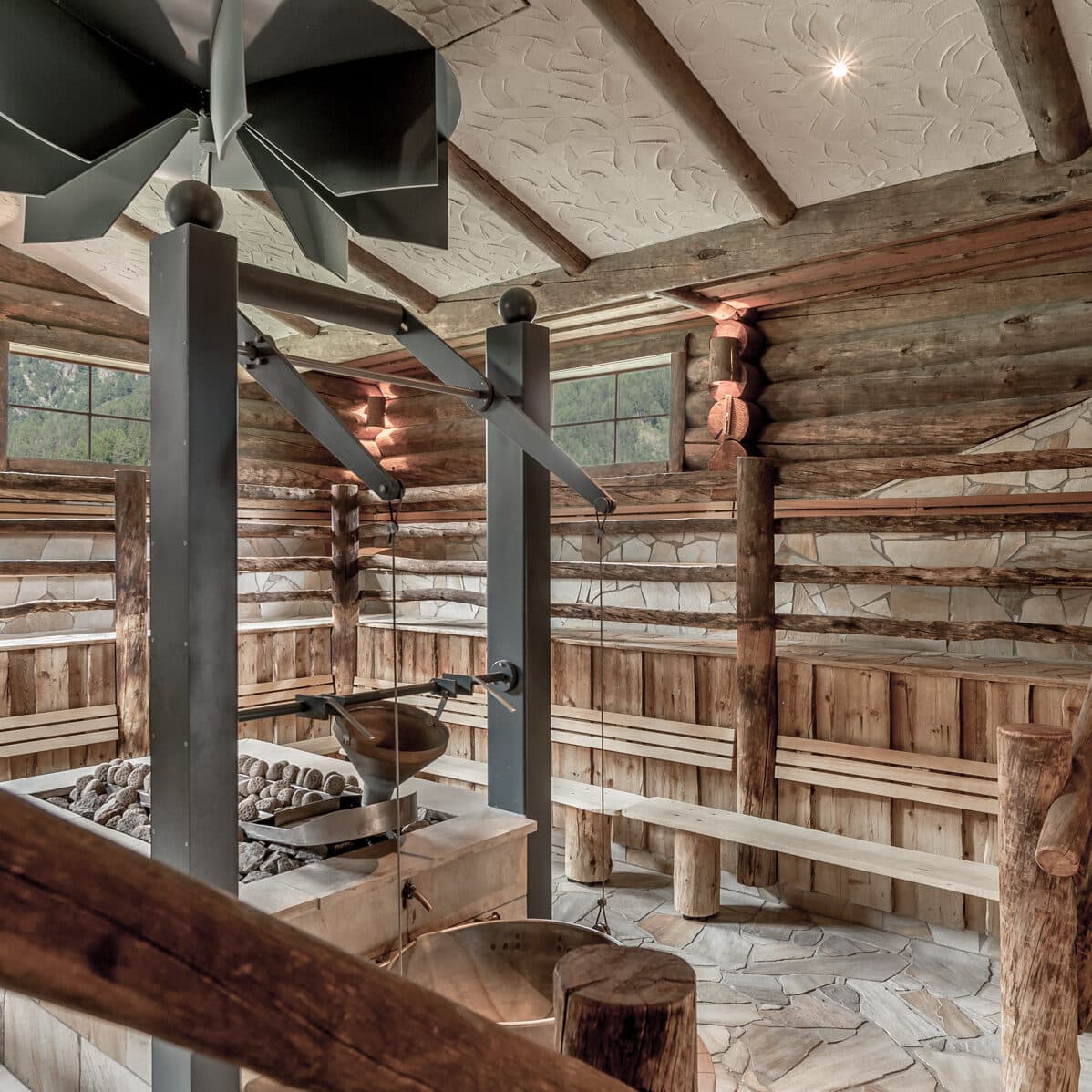 Ein rustikaler Saunaraum mit Holzbänken, Steinboden, Holzwänden und einem zentralen Ofen mit großen Metallvorrichtungen unter einer dekorativen Decke.