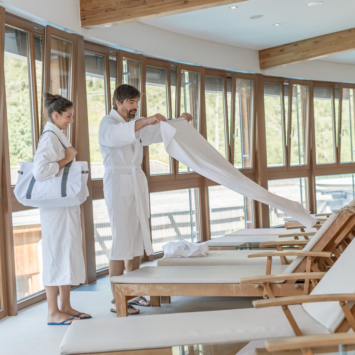 Zwei Personen in weißen Bademänteln bereiten Liegestühle mit Handtüchern in einem sonnenbeschienenen Wellnessbereich neben großen Fenstern vor.