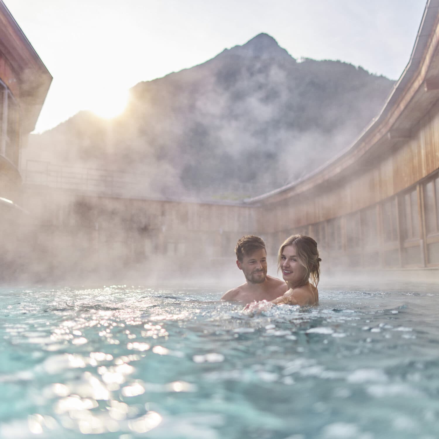 Ein Mann und eine Frau entspannen sich in einem Außenbecken mit heißen Quellen und aufsteigendem Dampf, umgeben von Holzwänden und Bergen im Hintergrund.