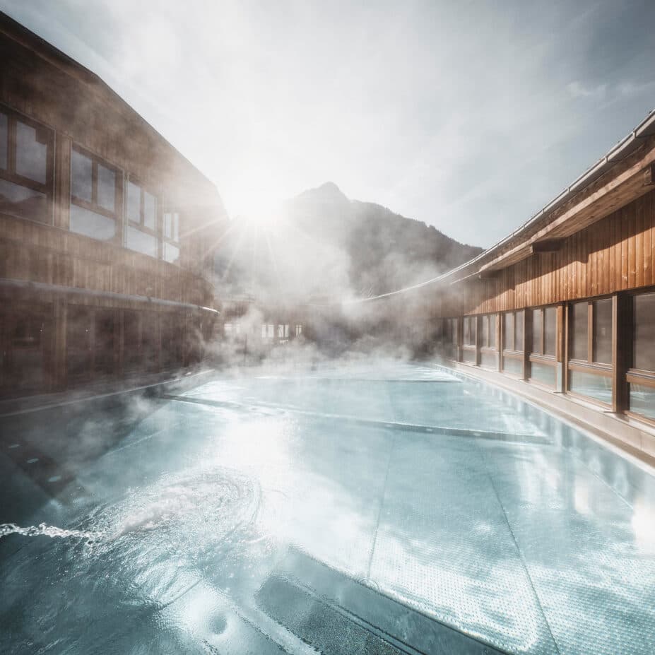Beheizter Außenpool, umgeben von Holzgebäuden, aus denen Wasserdampf aufsteigt, während im Hintergrund bei hellem Sonnenlicht die Berge zu sehen sind.