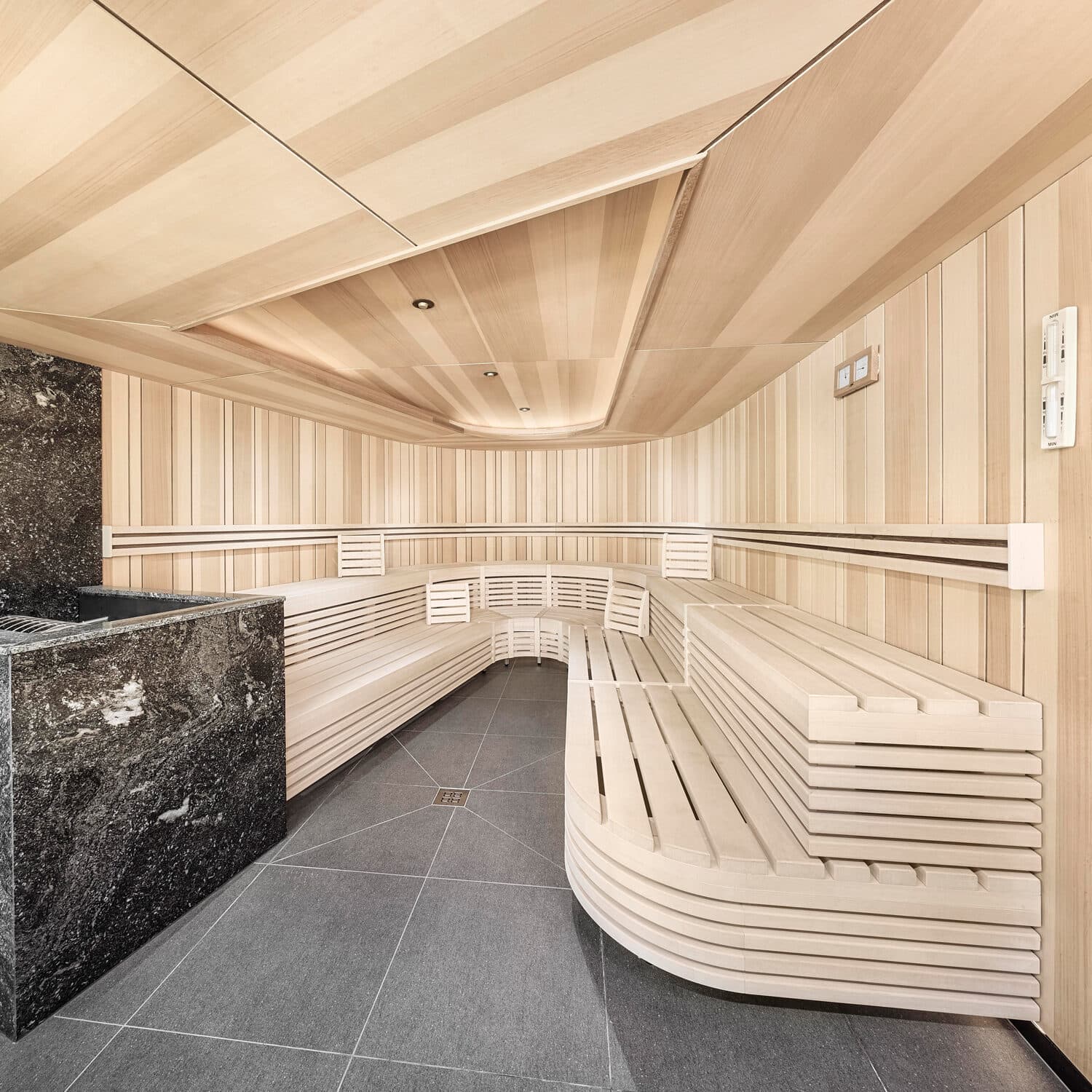 Moderne Holzsauna mit hellen Holzbänken, einem schwarzen Steinofen und gefliestem Boden. Der Raum ist leer und mit eingebauten Deckenleuchten gut beleuchtet.