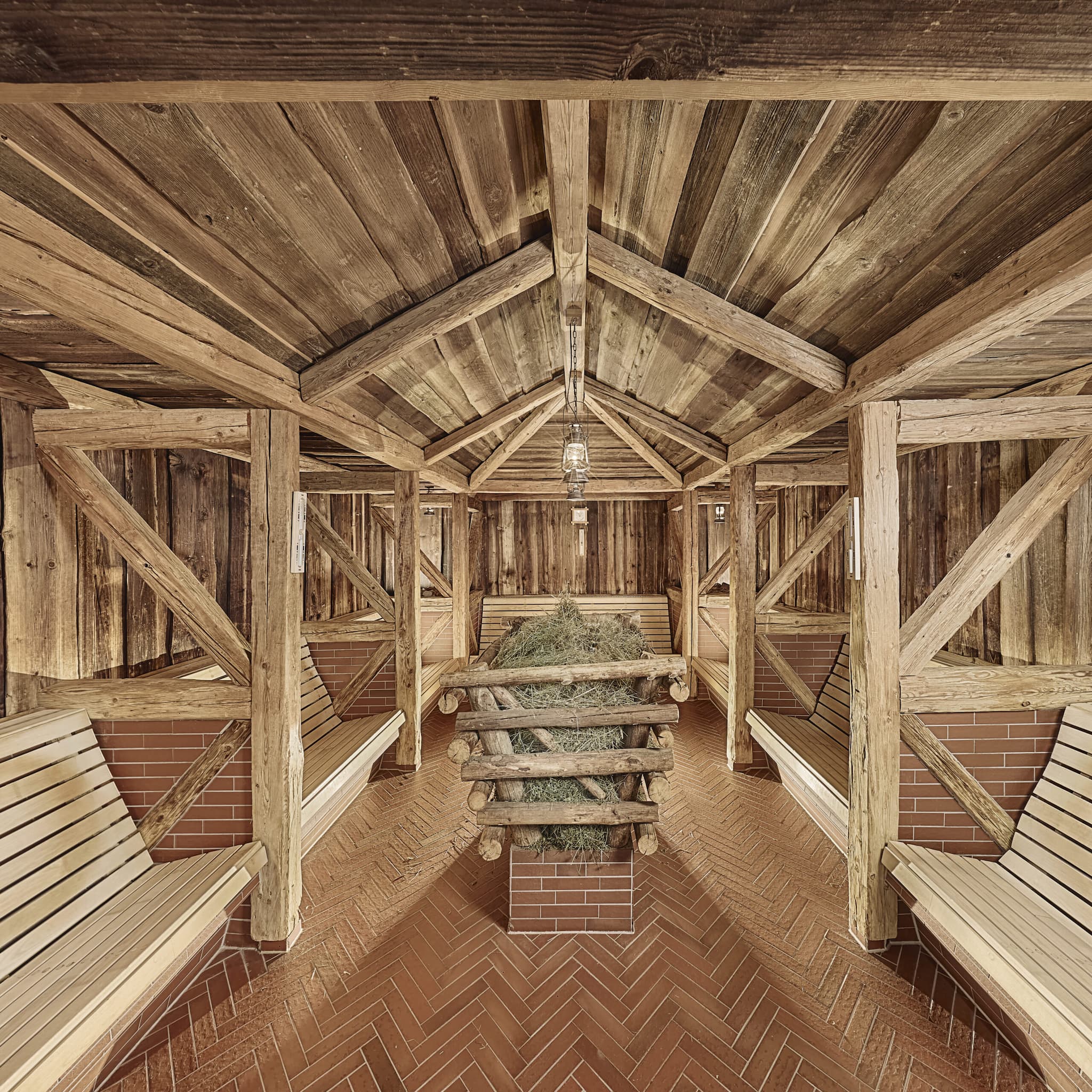 Das Innere einer rustikalen Holzsauna mit abgewinkelten Bänken an den Seiten, gemauertem Boden und einem zentralen, begrünten Blockhaus.