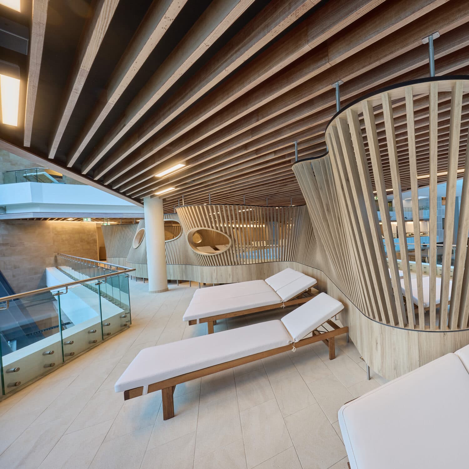 Moderner Indoor-Loungebereich mit Holzlattenarchitektur, weißen gepolsterten Lounge-Sesseln und Glasgeländer, der eine entspannte und zeitgemäße Atmosphäre schafft.