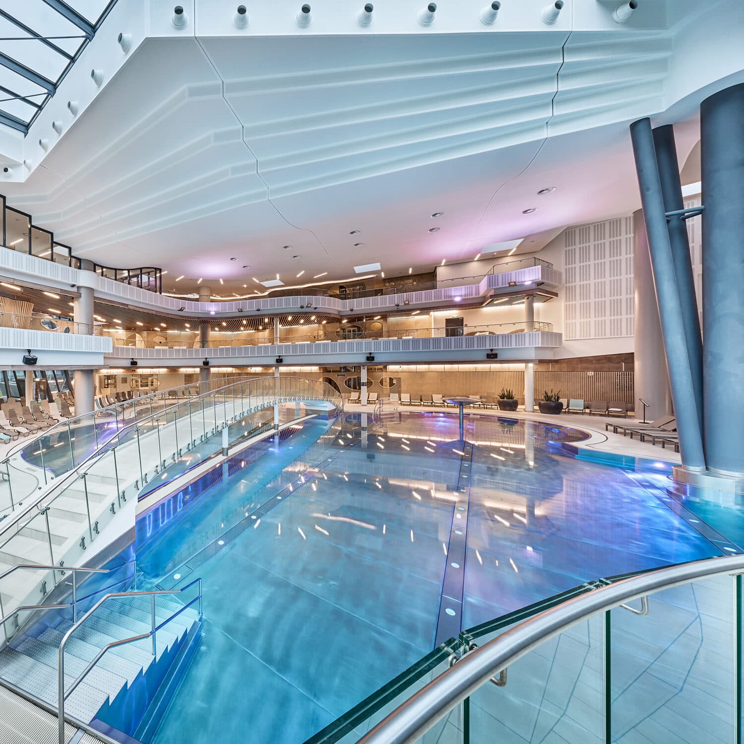 Hallenbad mit moderner Architektur, Glasgeländer und stimmungsvoller Beleuchtung; mehrere Balkone und Sitzbereiche mit Blick auf den Pool.