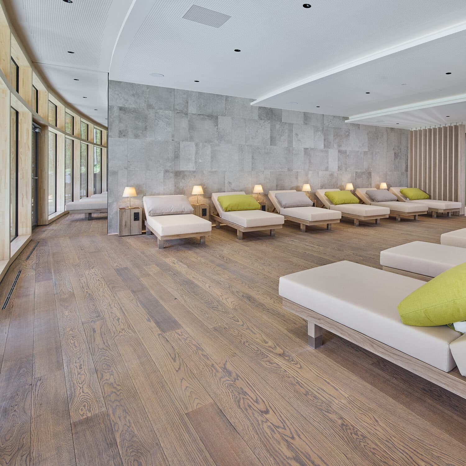 Moderner Entspannungsraum mit Holzboden, beigen Liegen, grünen Kissen, kleinen Lampen und großen Fenstern an einer Wand.