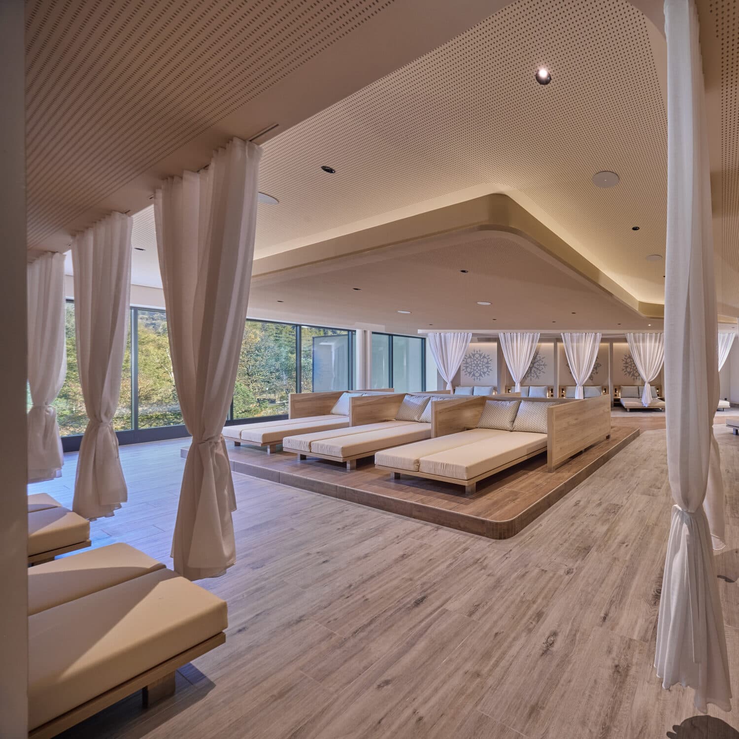 Geräumiger Spa-Ruheraum mit mehreren großen Podestbetten, weißen Vorhängen als Sichtschutz, Holzfußboden und großen Fenstern mit Blick ins Grüne.