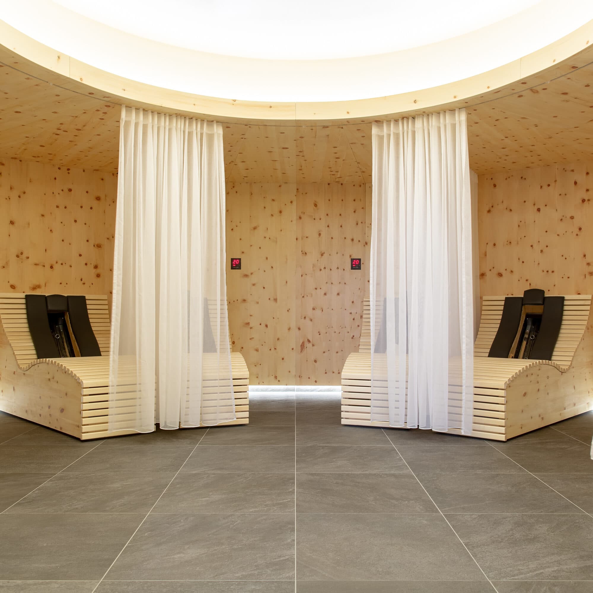 Zwei moderne Loungesessel aus Holz sind durch weiße Vorhänge in einem spa-ähnlichen Raum mit grau gefliesten Böden und hellen Holzwänden und -decken voneinander getrennt.
