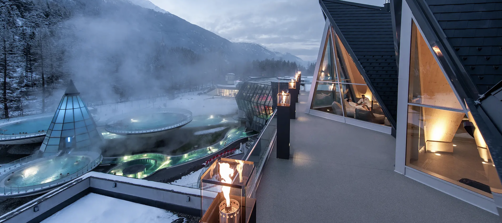 Ein schneebedecktes Bergresort mit dampfenden Außenpools, modernen Glasstrukturen und einem Balkon mit Feuerlaternen in der Abenddämmerung.