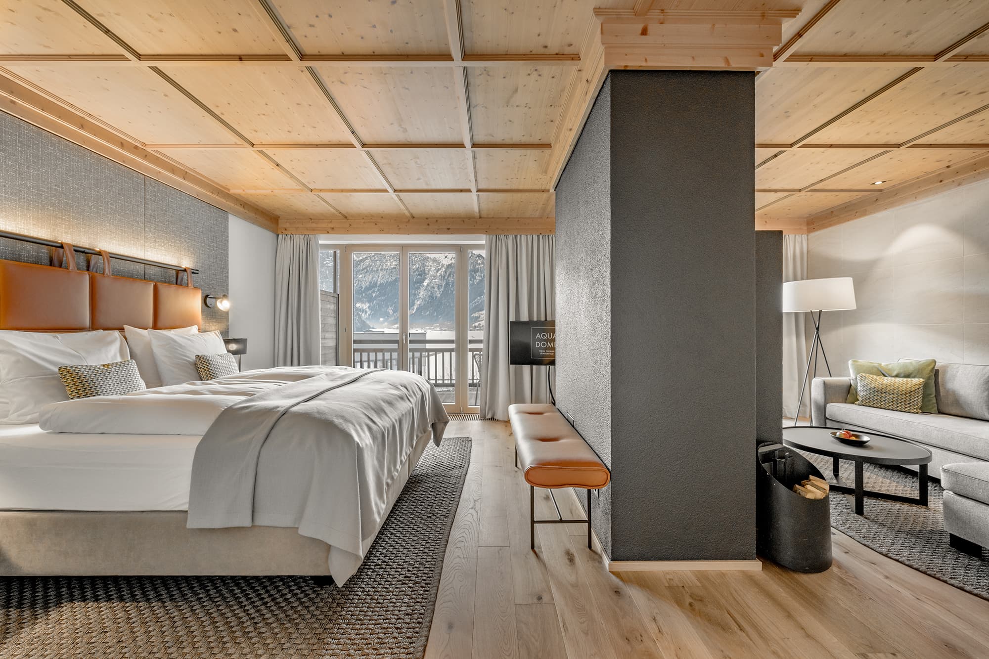 Modernes Hotelzimmer mit großem Bett, Sofa und Sitzbank; holzgetäfelte Decke und Boden; Blick auf die Berge durch Glastüren, die auf einen Balkon führen.