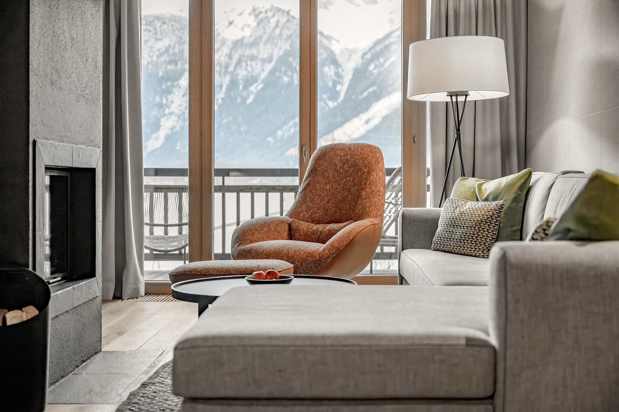 Ein modernes Wohnzimmer mit einem grauen Sofa, einem orangefarbenen Sessel, einer Stehlampe und einem Kamin, mit großen Fenstern mit Blick auf verschneite Berge und einen Balkon.