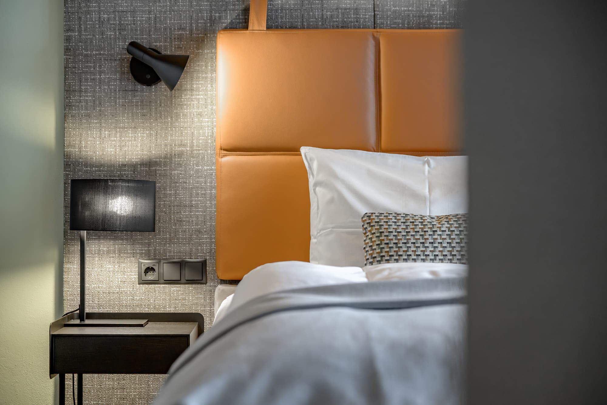 Ein modernes Hotelzimmer mit einem Kopfteil aus braunem Leder, einem gemachten Bett mit weißer Bettwäsche, einem Nachttisch und zwei Lampen vor einer strukturierten grauen Wand.
