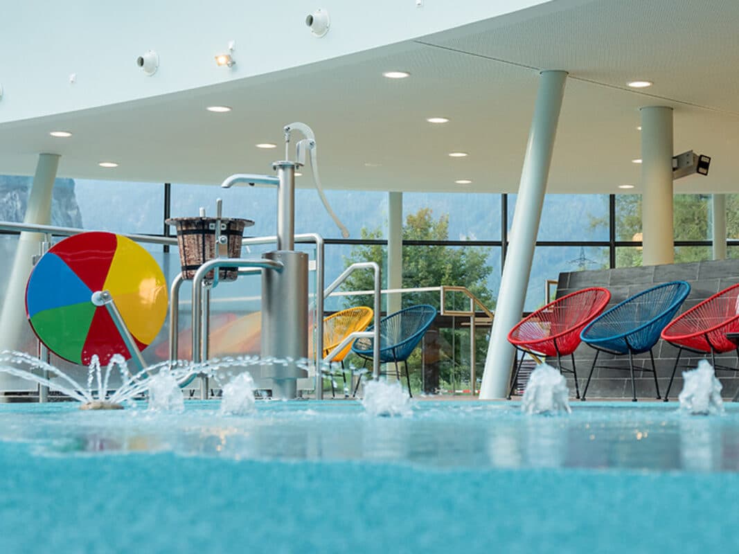 Innenpool mit Wasserfontänen, bunten Liegestühlen, einem großen bunten Rad und großen Fenstern mit Blick auf die Bäume und Berge draußen.