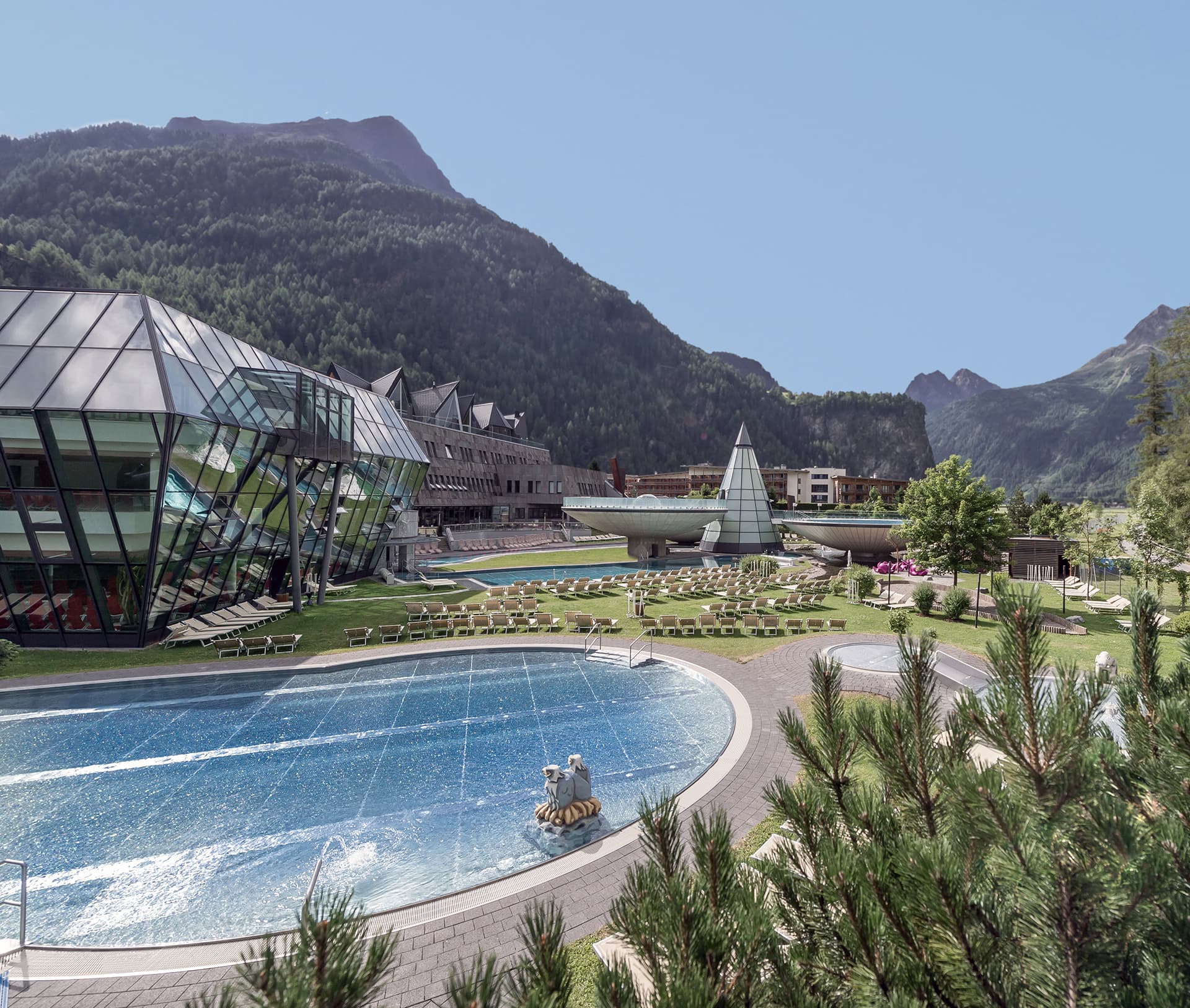 Glasgebäude und ein Außenpool mit Liegestühlen bieten vor der Kulisse grüner Berge und eines klaren Himmels einen herrlichen Anblick.