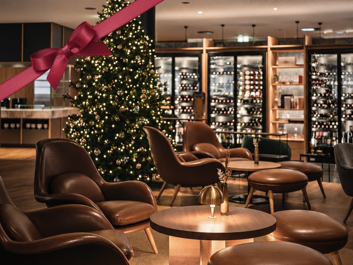 Eine moderne Lounge mit Ledersesseln, runden Tischen, einem geschmückten Weihnachtsbaum und Regalen voller Weinflaschen – ideal für festliche Zusammenkünfte oder Besprechungen. In der oberen linken Ecke ist ein rotes Band mit einer Schleife zu sehen.
