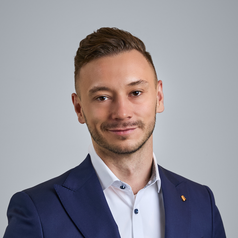 Ein Mann in einem blauen Anzug und einem weißen Hemd, der vor einem grauen Hintergrund lächelt.